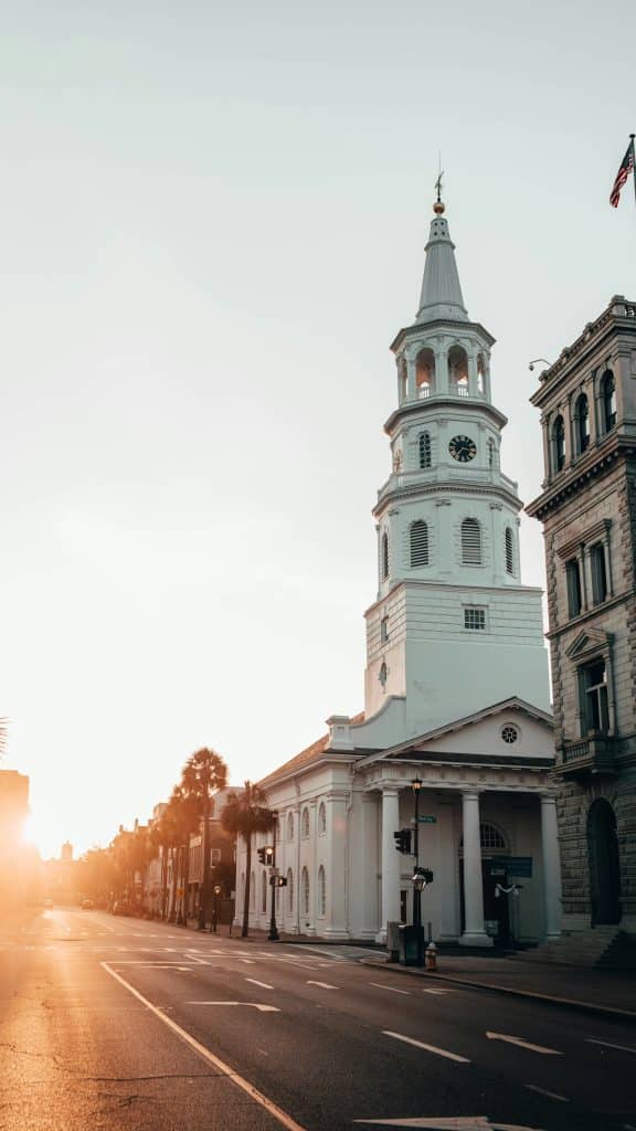 St. Philip's Church steeple in Charleston, SC, rises above Meeting Street at sunrise, with the American flag and historic buildings lining the quiet road.